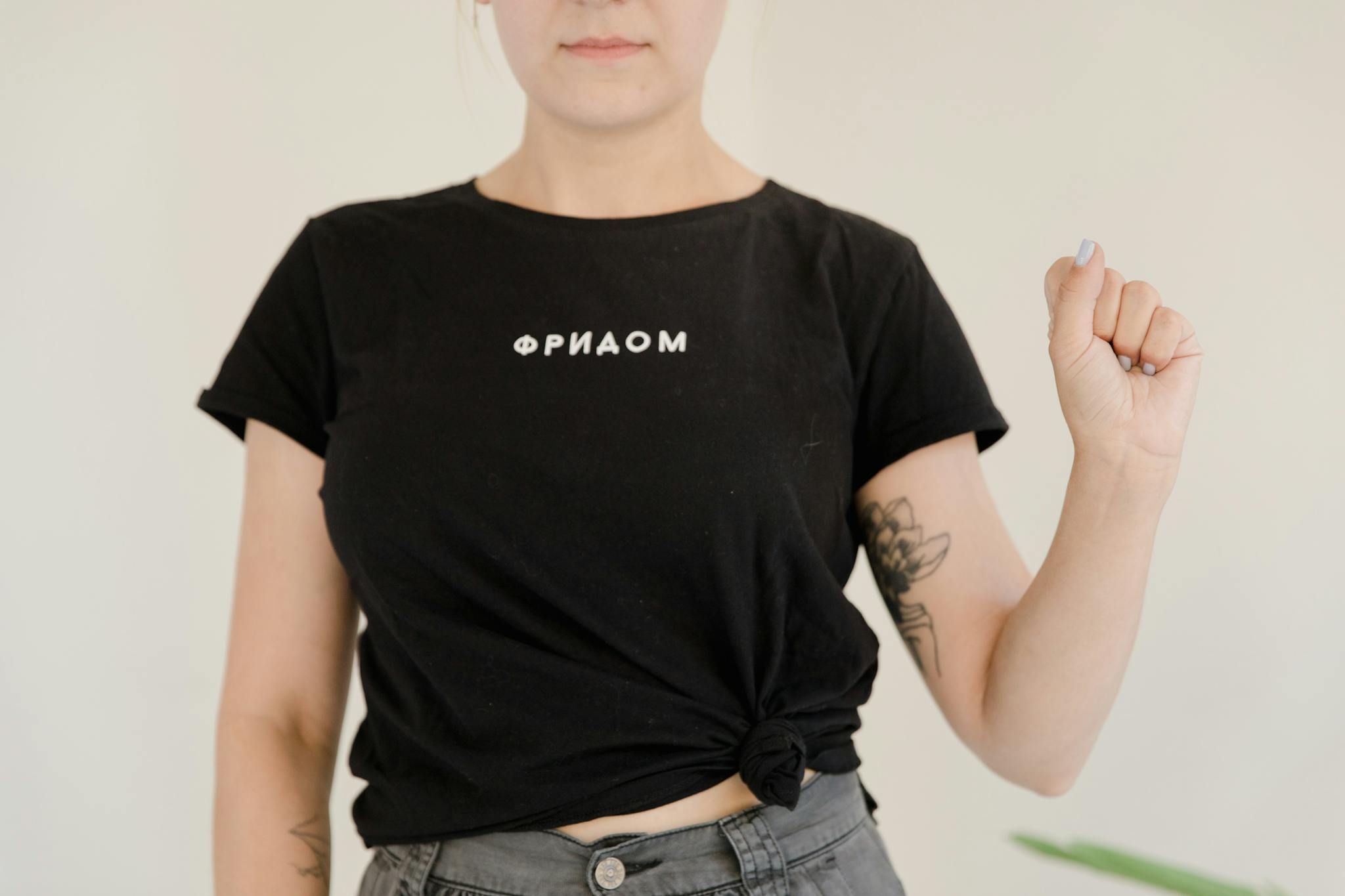 Woman wearing a black T-shirt with "ФРИДОМ" print and floral tattoos on arm indoors.